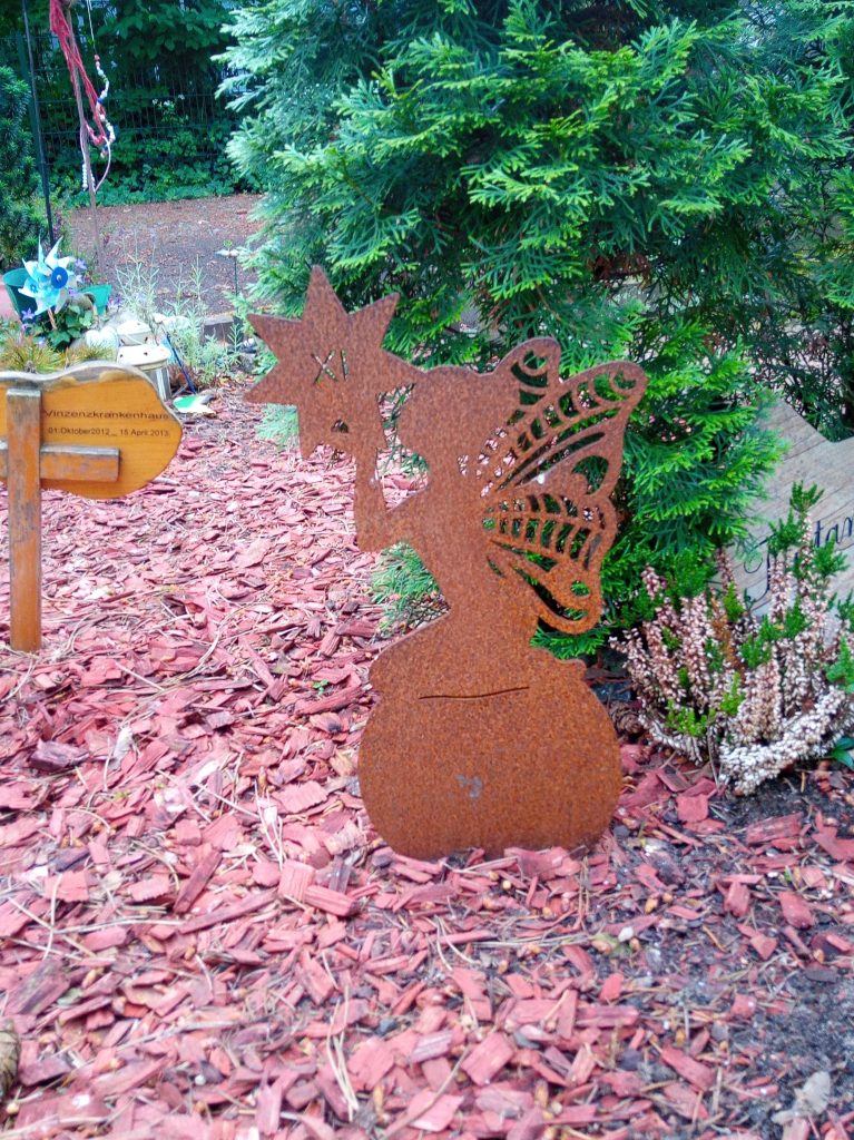 Foto einer Schmetterlingselfe aus Metall als Nahaufnahme; im Hintergrund ein grüner Busch, daneben ein kleines Holzkreuz mit Schild zur Markierung welche Gemeinschaftsbestattung hier stattgefunden hat.